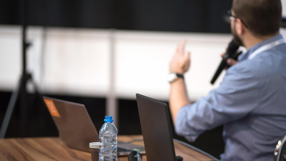 A lecturer sits at a desk with a microphone.