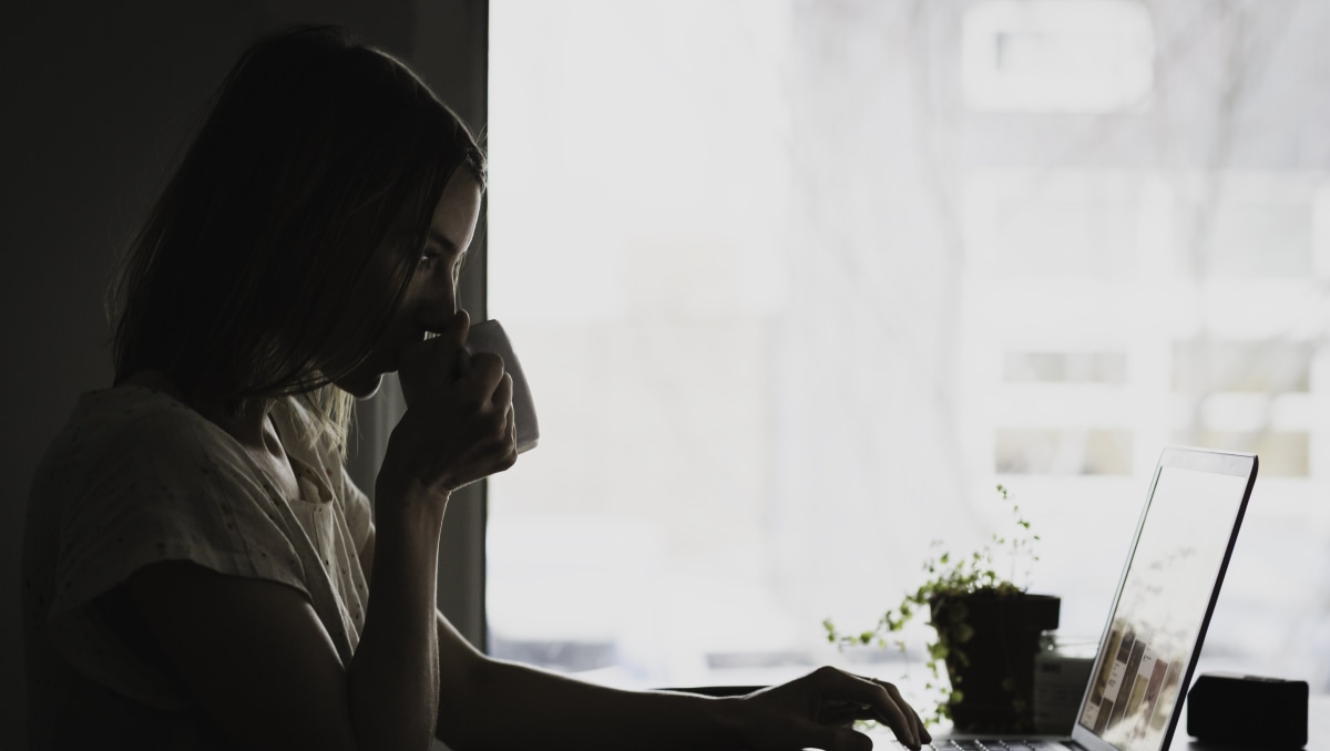 A woman sitting in front of a laptop in a dark room, drinking from a mug.